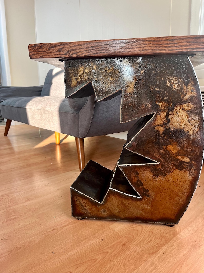K&ouml;nnte beinhalten: Eine rustikale Holztischplatte mit einem einzigartigen, abstrakten Metallfu&szlig;. Der Fu&szlig; besteht aus mehreren zusammengeschwei&szlig;ten Metallteilen und hat eine rostige Oberfl&auml;che.