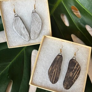 May include: Two pairs of handmade earrings displayed in beige boxes. One pair is light gray with a dark border, the other is dark brown with etched lines. Both pairs have gold-colored ear wires and are set against a green leaf background.