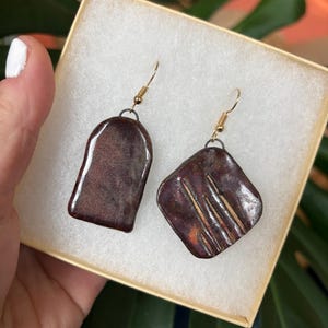 May include: A pair of handmade ceramic earrings in a gift box. One earring is a rounded rectangle, the other is a square with diagonal lines. Both are glazed in a deep burgundy color with gold-colored ear wires.