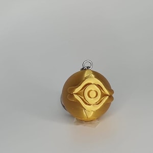 Puede incluir: Un colgante dorado con un diseño detallado de ojo. El colgante es esférico con un lazo plateado en la parte superior para sujetarlo a una cadena o llavero. El diseño del ojo es una característica destacada, con líneas y curvas intrincadas.