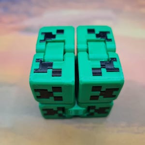 Può includere: Un giocattolo fidget verde e nero, con un design Creeper. Il giocattolo è composto da diversi blocchi collegati, ognuno con una faccia Creeper. I blocchi sono verdi con dettagli neri pixelati. Lo sfondo è un mix sfocato di colori.