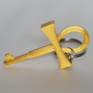 Pode incluir: Um porta-chaves dourado em forma de ankh. O ankh, um símbolo da vida, está preso a um porta-chaves prateado. O ankh tem uma superfície texturizada e é feito de um material sólido. O porta-chaves está sobre um fundo neutro.