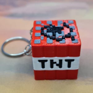 May include: A red, white, and black TNT block keychain. The block features the letters "TNT" in black on a white band. The top of the block has a pixelated design in black and gray. A silver key ring is attached.