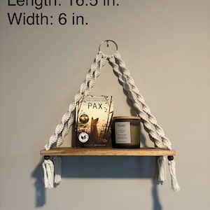 以下が含まれることがあります： 本とキャンドルを置いた木製の棚が付いた、マクラメの吊り下げ棚。棚の高さは45.7cm、長さは41.9cm、幅は15.2cmです。棚は白いマクラメロープで吊り下げられています。