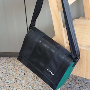 Könnte beinhalten: Schwarze Messenger-Tasche aus recycelten Materialien mit einem strukturierten Reifen-Design. Die Tasche hat einen grünen Boden und einen schwarzen Gurt. Der Markenname "mnmur" ist auf der Vorderseite sichtbar.