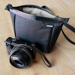 May include: Black camera bag with an open zipper, next to a black camera with a lens and strap. The bag has a rectangular shape and a small tag that reads "mnmur". The camera strap is black and tan.