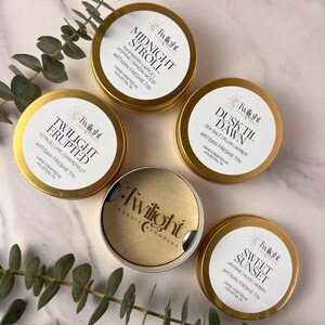 May include: Four gold candle tins with labels and names of scents. The scents are Midnight Stroll, Dusk Til Dawn, Twilight Erupted, and Sweet Sunset. Each tin has a gold lid and a white label with black text. The candles are handcrafted in Houston, Texas.