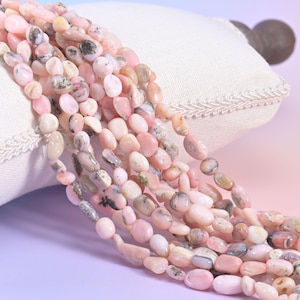 Puede incluir: Hebras de cuentas de piedras preciosas de ópalo rosa. Las cuentas de forma ovalada varían en tonos de rosa, algunas con inclusiones grises y marrones. Las cuentas se exhiben sobre una almohada blanca con un borde decorativo.