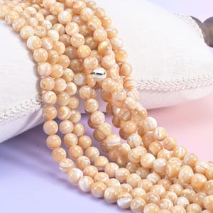 May include: Strands of round, light peach-colored beads with white marbling. The beads are arranged in multiple strands, creating a full and textured appearance. The beads are likely used for jewelry making.