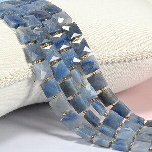 May include: Three strands of faceted, square-shaped blue gemstone beads arranged in parallel rows. Each bead features a polished, multi-faceted surface with varying shades of blue and grey, separated by small, round, metallic gold-colored spacer beads.