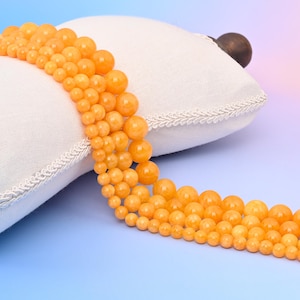 May include: Strands of orange gemstone beads in varying sizes are arranged on a white pillow with a decorative trim. The beads have a smooth, polished surface and a warm, amber hue, creating a gradient effect.