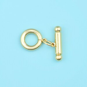 Op de afbeelding: Een goudkleurige toggle sluiting, bestaande uit een cirkelvormige ring en een staaf met afgeronde uiteinden, verbonden door kleine schakels. De sluiting staat tegen een lichtblauwe achtergrond. Geschikt voor kettingen en armbanden.