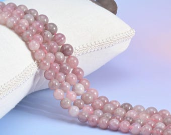 May include: Strands of pink and mauve gemstone beads are draped over a white pillow with a decorative edge. The beads are round and vary in shade, creating a gradient effect. The background is a soft gradient of blue and pink.