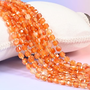 May include: Strands of orange gemstone beads with a faceted, square shape. The beads are strung together with small gold-colored spacers. The beads are resting on a white pillow with a lace trim.