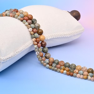 May include: A strand of round, multi-colored, natural stone beads. The beads are a mix of brown, green, and white with a speckled pattern. The beads are strung on a white cord and are displayed on a white pillow.