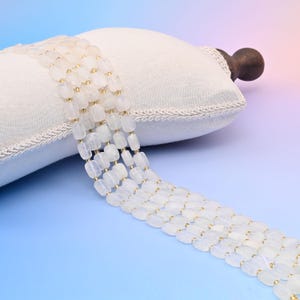 May include: A strand of white moonstone beads with a rectangular shape. The beads are strung on a gold wire.