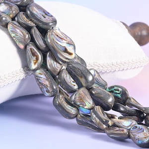 May include: A strand of irregular, tumbled abalone shell beads featuring a dark, charcoal-colored base with iridescent swirls of green, blue, and purple. The beads are polished to a smooth finish, showcasing the natural, multi-colored luster.
