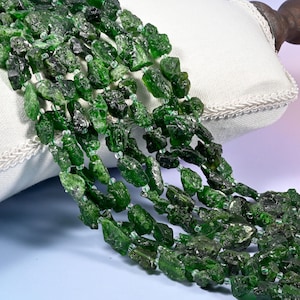 May include: A multi-strand necklace featuring raw, deep green, irregularly shaped gemstone nuggets. The stones are strung together with small, clear, spherical spacer beads, creating a textured, layered appearance with a vibrant, natural aesthetic.
