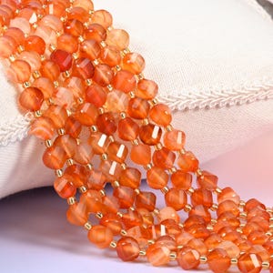 May include: A multi-strand necklace featuring faceted, translucent orange carnelian beads. The beads are arranged in several parallel rows, separated by small, metallic gold-colored spacer beads, creating a vibrant and textured appearance.