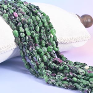 May include: Strands of polished gemstone beads in shades of green and maroon. The beads are oval-shaped and have a natural, organic appearance. The beads are displayed on a white pillow.