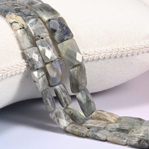May include: A strand of faceted rectangular labradorite beads featuring a mix of gray, blue, and cream tones with natural inclusions and a polished finish. The beads are arranged in a vertical line, showcasing their unique mineral patterns and light-reflecting surfaces.