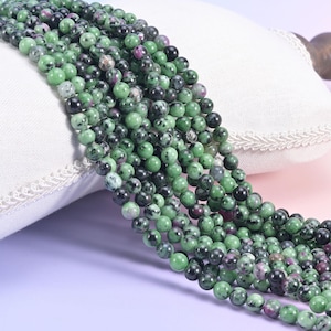 May include: Strands of round gemstone beads in shades of green, black, and hints of red. The beads are smooth and appear to be natural stone, likely for jewelry making. The beads are resting on a white pillow.