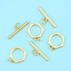 May include: Gold-colored jewelry clasps, including toggle bars and hexagonal rings, are scattered on a light blue surface. The clasps have a shiny finish and are designed for jewelry making. The toggle bars have a loop at each end for attachment.