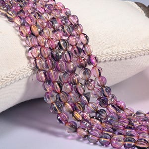 May include: A strand of translucent, round, ribbed glass beads in shades of purple and clear. Each bead features vertical grooves that catch the light, creating a shimmering effect. The beads are strung together in a continuous, multi-row arrangement.