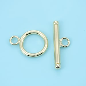 May include: A gold-colored toggle clasp set against a light blue background. The set includes a circular ring with a small loop and a straight bar with a loop and ball ends. Ideal for jewelry making.