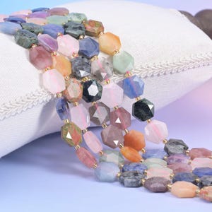 May include: A multi-strand necklace featuring faceted gemstone beads in various colors, including pink, blue, green, and orange. The beads are strung together with gold-colored accents, creating a colorful and eye-catching accessory.