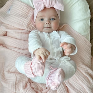 May include: A baby in a white ribbed jumpsuit with a front zip, pink knitted booties, and a large pink bow headband. The baby is on a pink blanket and a white pillow.