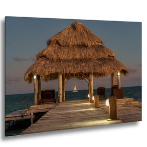 May include: A wooden pier extends into the ocean, leading to a thatched-roof gazebo with lights. The gazebo has wooden posts and a layered roof. A sailboat is visible in the distance, under a dusky sky. The scene evokes a tropical, relaxing atmosphere.