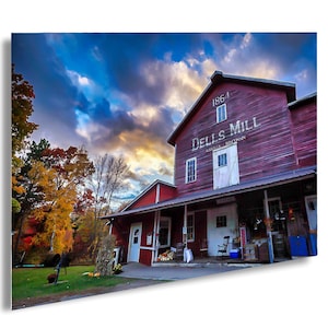 May include: A red wooden building with a sign that reads "Dell's Mill Augusta Wisconsin 1864". The building has a porch with a rocking chair and a few potted plants. The sky is blue with white clouds.