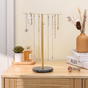 May include: A gold metal jewelry stand with a marble base. The stand has multiple hooks for hanging necklaces and bracelets. The stand is on a wooden surface.