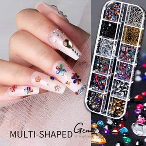 Op de afbeelding: Een hand met lange, versierde nagels, met diverse multi-vormige edelstenen en nail art. De nagels zijn versierd met strass, bloemen, sterren en andere kleurrijke versieringen. Een doorzichtige plastic container gevuld met diverse nail art edelstenen is ook zichtbaar.