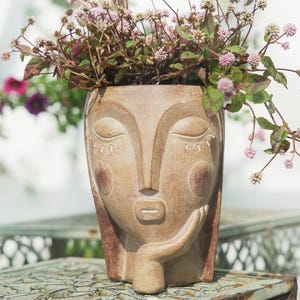 May include: A beige-colored, face-shaped planter with closed eyes and a hand resting on the chin. The planter is filled with pink and green flowering plants. The planter has a distressed finish and is sitting on a metal surface.