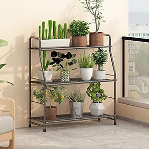 May include: A three-tiered, dark gray metal plant stand holding various potted plants. The plants include cacti, herbs, and small trees. The stand is on a light gray surface, with a balcony and building visible in the background.