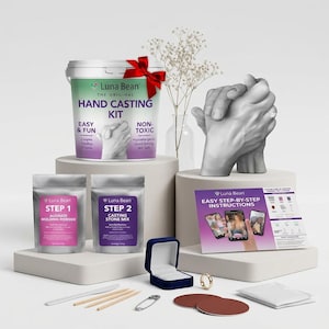 May include: A Luna Bean hand casting kit featuring a white bucket with a red bow, two silver pouches labeled Step 1 and Step 2, a set of instructions, a blue ring box, a gold ring, a plastic knife, wooden sticks, and a finished gray hand sculpture.