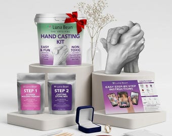 May include: A Luna Bean hand casting kit featuring a white bucket with a red bow, two silver pouches labeled Step 1 and Step 2, a set of instructions, a blue ring box, a gold ring, a plastic knife, wooden sticks, and a finished gray hand sculpture.