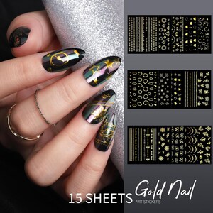 Op de afbeelding: Zwarte nagellak met gouden nail art stickers. De stickers hebben verschillende ontwerpen, waaronder sterren, lijnen en geometrische vormen. De afbeelding toont ook een hand met een zilveren ring en de tekst "15 SHEETS Gold Nail ART STICKERS."
