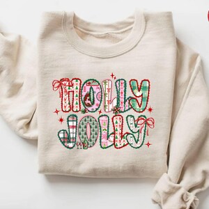 May include: A sand-coloured sweatshirt featuring the words "HOLLY JOLLY" in a festive design. The letters are decorated with plaid patterns, Christmas trees, and bows. Red stars and accents enhance the holiday theme.