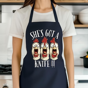 May include: Navy blue apron with the text "SHE'S GOT A KNIFE !!" above a cartoon image of three chickens with wide eyes and open beaks. The apron has a bib and neck strap.