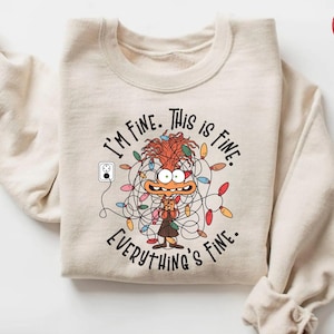 Może przedstawiać: Beżowa bluza z napisem "I'M FINE. THIS IS FINE. EVERYTHING'S FINE." Postać z kreskówki jest zaplątana w lampki choinkowe. Przedstawiono również gniazdko elektryczne. Bluza ma długie rękawy.