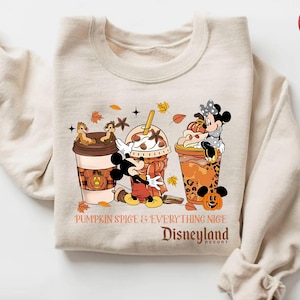 Peut inclure: Sweat-shirt couleur sable avec un motif coloré de Mickey et Minnie Mouse, Chip et Dale, et trois boissons sur le thème de l'automne. Le texte indique "Pumpkin Spice & Everything Nice Disneyland Resort."