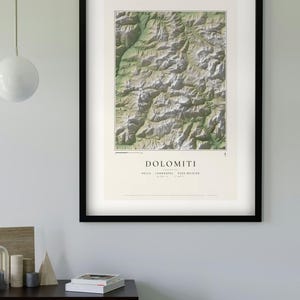 以下が含まれることがあります： 緑の谷と灰色の山頂が描かれたドロミテ山脈の地形図の額入りプリント。テキストには「DOLOMITI」「SELLA」「LANGKOFEL」「PUEZ-GEISLER」と記載され、地理座標が含まれています。プリントは黒いフレームに入っています。