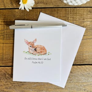 May include: A white greeting card featuring a watercolor illustration of a brown fawn resting on green grass. Below the fawn, the text reads: Be still & know that I am God. Psalm 46:10. A silver pen rests horizontally across the top of the card.
