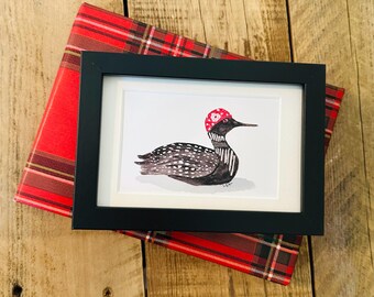 May include: A framed watercolor print of a loon wearing a red and white polka dot hat. The black framed artwork rests on a red and black plaid patterned wrapping paper. The background is a wooden surface.