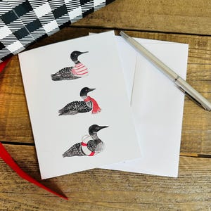 May include: A white greeting card featuring three illustrations of loons. The top loon wears a red and white striped sweater, the middle loon wears a red scarf, and the bottom loon wears a red and white life preserver. A silver pen rests nearby.