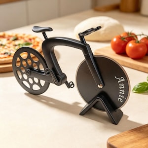 May include: A black bicycle-shaped pizza cutter with a circular blade featuring the name Annie engraved in white script. The cutter has a rear wheel with a perforated design and a front wheel that serves as the blade, supported by a black stand.