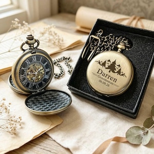 Puede incluir: Un reloj de bolsillo de tono latón con esfera negra y números romanos, con la tapa abierta mostrando los engranajes. Un segundo reloj a juego está en una caja negra, grabado con un bosque de montaña, el nombre Darren y Groomsman 08.09.26.